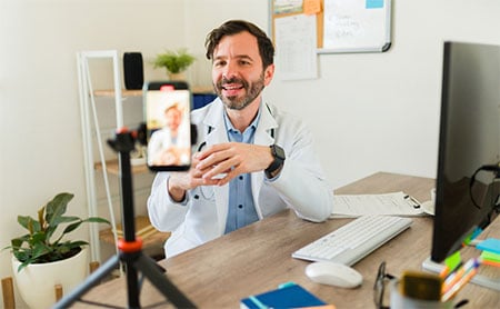 A person in a white lab coat sits at a desk with a computer, engaging in a video call.