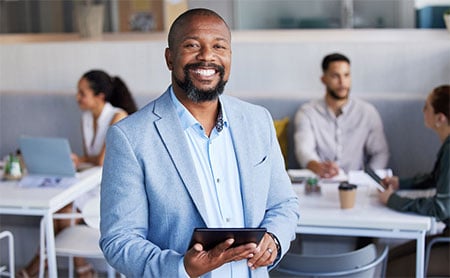 Smiling man in a light blue blazer holding a tablet in an office setting with colleagues in the background.