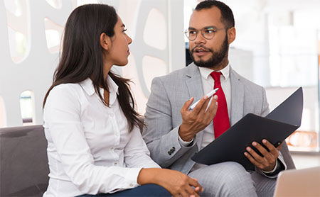 Two colleagues seated on a couch in a discussion, with man holding a folder.