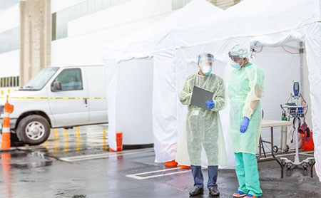 Two healthcare workers in protective gear outside a white tent.