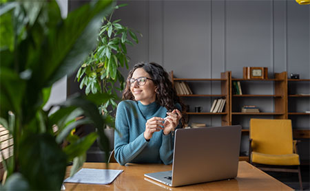 Woman with glasses sitting at a desk with a laptop smiling, surrounded by a plant and bookshelves.